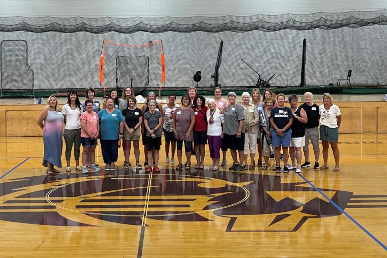 Female student-athlete alumni gathered in 2022 for a reunion with Coach Kim Fischer. Fischer is in the front row wearing a gray shirt and red shoes.